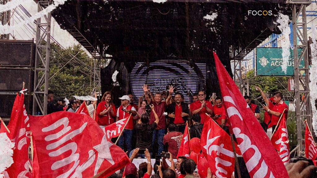 Fotografía del mitin de inicio de campaña electoral del partido FMLN. En un tarima, al centro, se encuentra el candidato presidencial Manuel 'El Chino' Flores y los dirigentes del partido. Abajo, los militantes alzan banderas del partido.